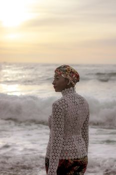 A woman stands by the ocean in a headscarf during a beautiful golden sunset.