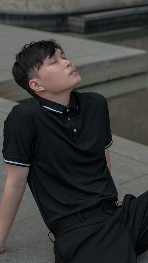 Free A young man in a black polo shirt sits outdoors, eyes closed in relaxation. Stock Photo