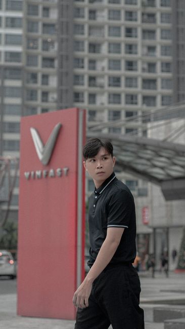 Young man in a black polo shirt walking in an urban street with modern architecture in the background.