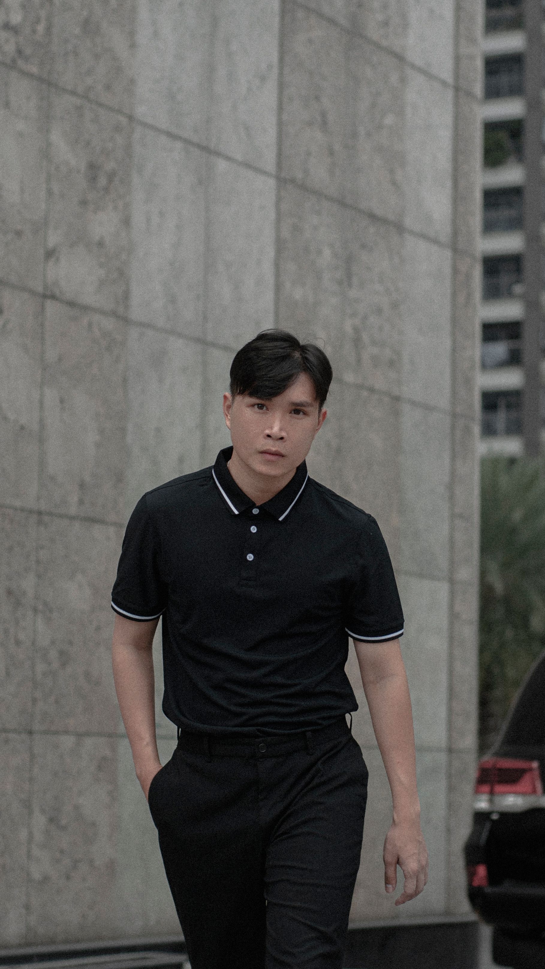 Homem asiático caminhando com confiança em ambiente urbano, vestido com camisa polo preta e calças.