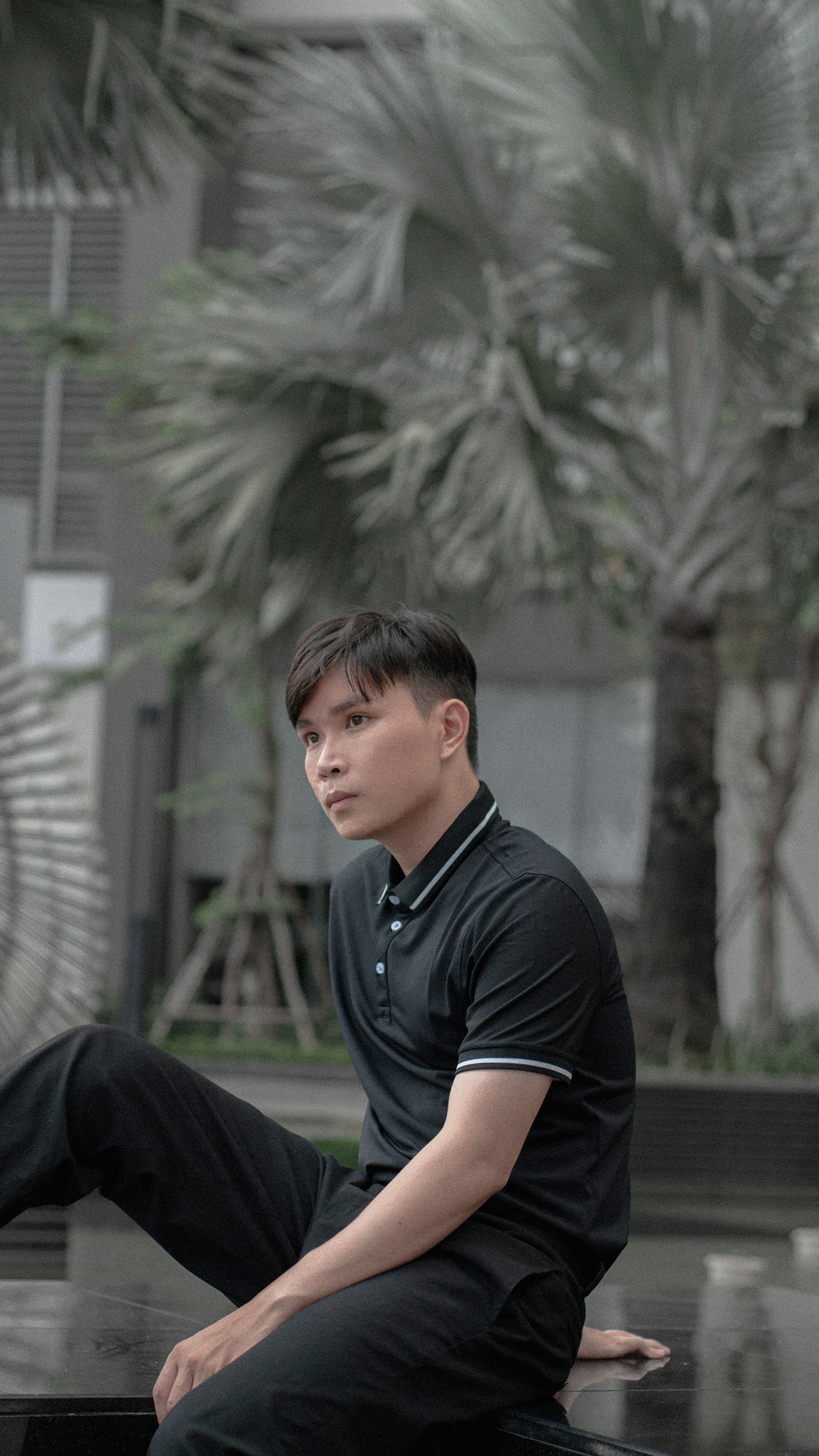 Young man in a black polo shirt sitting outdoors in an urban environment.