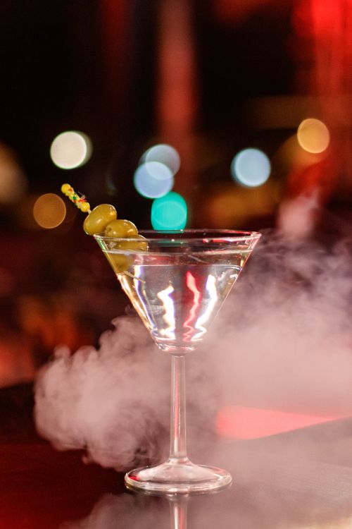 Free A classic martini garnished with olives in a smoky, atmospheric setting with bokeh lighting. Stock Photo