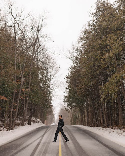 A Woman in Black Outfit Crossing the Road Between Tall Trees
