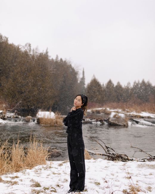 Woman in black outfit posing by a snowy riverbank during snowfall.