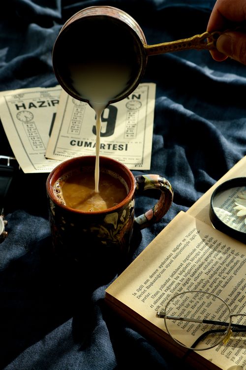 Free A cozy indoor scene of milk being poured into a coffee cup, with an open book and eyeglasses on a dark cloth background. Stock Photo