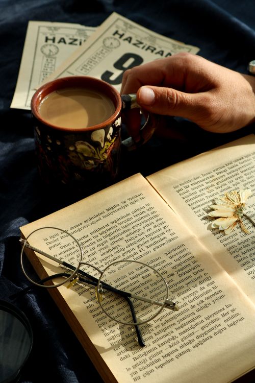 Free A warm and inviting image of a hand holding a coffee cup next to an open book and glasses. Stock Photo