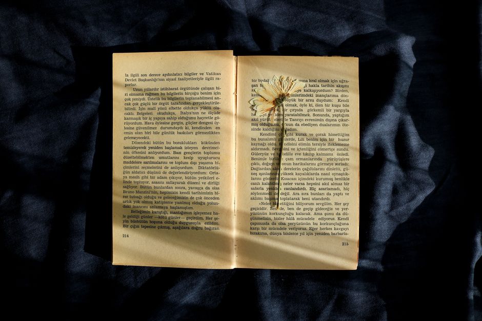 An open book with a dried flower placed on a dark fabric background, in natural light.