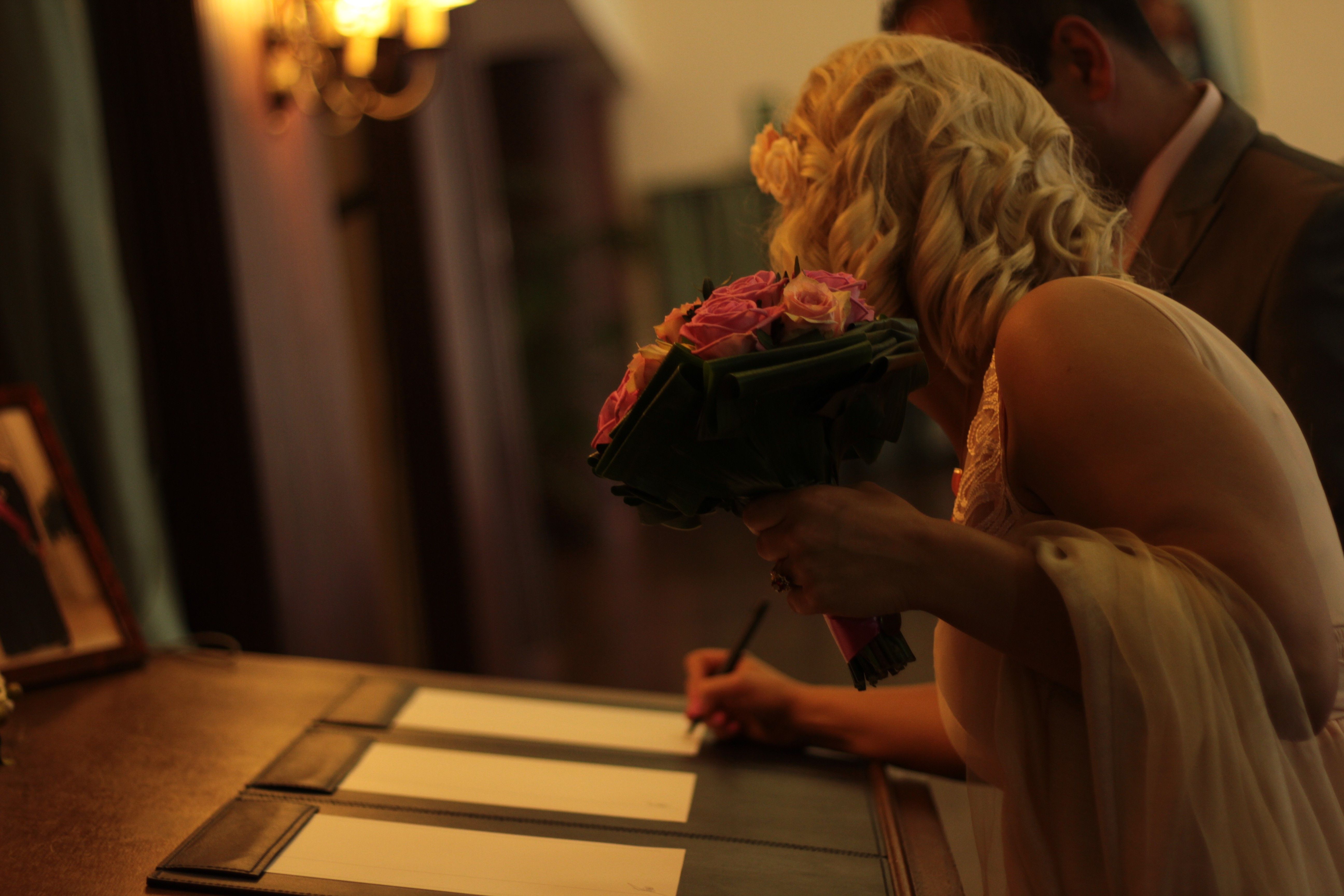 A bride signing a document indoors, holding a bouquet of roses during a wedding ceremony.