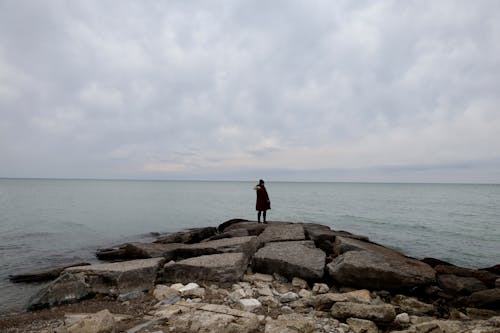 Woman in a Coat Standing on a Shore with Rocks