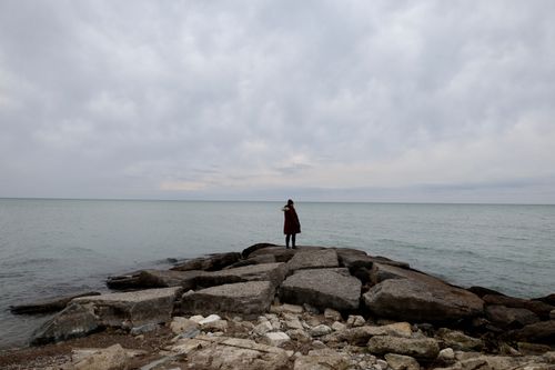 Free A solitary woman stands on rocks by a calm sea under an overcast sky. Stock Photo