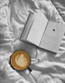 Crisp latte with beautiful foam art beside an open book on a cozy bed.