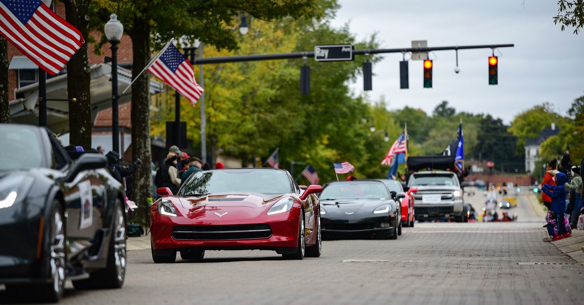 A vibrant parade in downtown Fayetteville, NC, featuring Corvettes and American flags.