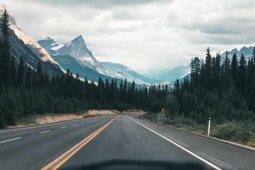 Empty highway through the majestic Canadian Rockies under a cloudy sky, framed by dense forest.
