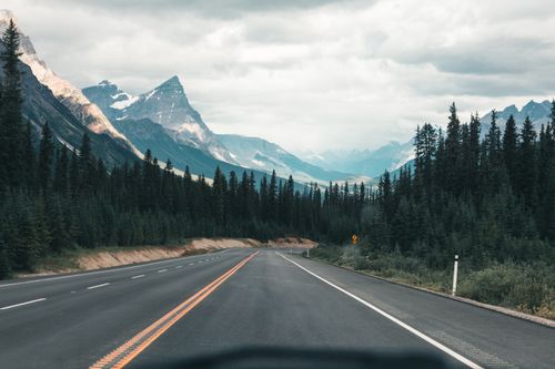 Free Empty highway through the majestic Canadian Rockies under a cloudy sky, framed by dense forest. Stock Photo