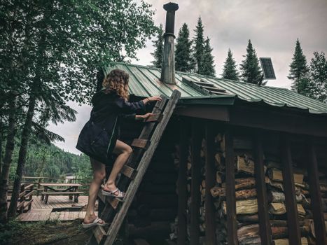 A woman climbs a wooden ladder to the roof of a rustic cabin surrounded by lush trees in Canada.