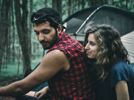 A young couple enjoys a moment outdoors at a scenic forest campsite with tents in the background.
