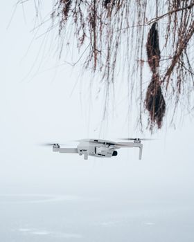 A white drone flies near leafless willow branches against a snowy winter backdrop, showcasing aerial technology.