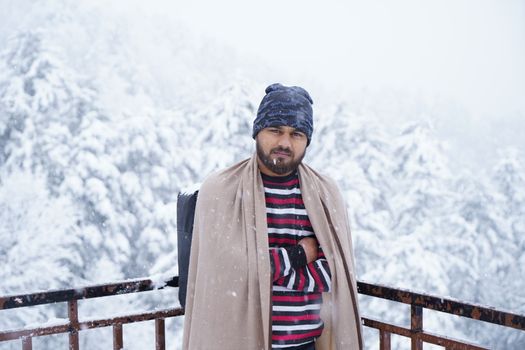 Man bundled in winter clothing standing amidst snowfall and snowy forest backdrop.