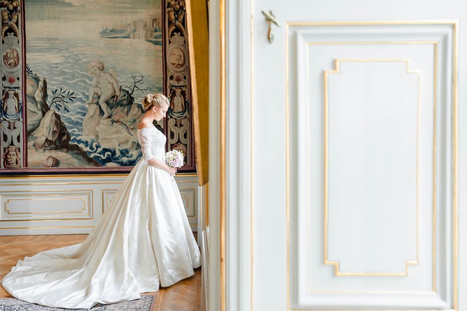 Graceful bride in white gown holding a bouquet, standing in a decorated room with classic architecture.