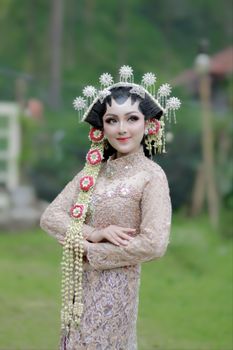 Elegant Indonesian bride in traditional attire with ornate headdress and nature backdrop.