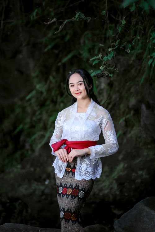 Free Asian woman in white lace top and floral skirt with red belt standing outdoors, smiling gently. Stock Photo