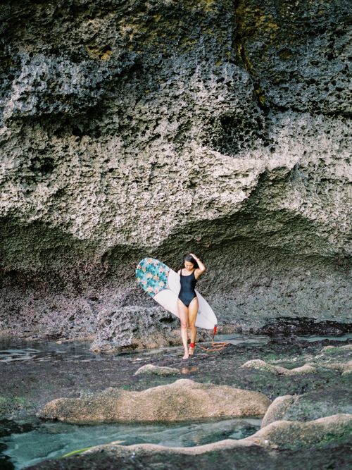 Free A woman with a surfboard stands by rocky cliffs in Bali, showcasing beach adventure. Stock Photo