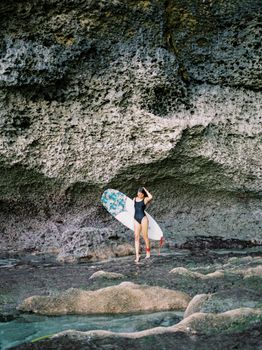 A woman with a surfboard under a dramatic rock formation on a Bali beach.