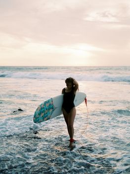 Woman surfer at Bali beach holding surfboard during sunset, showcasing beach lifestyle.