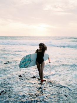 Woman with surfboard walks on a Bali beach at sunset, enjoying the ocean breeze.