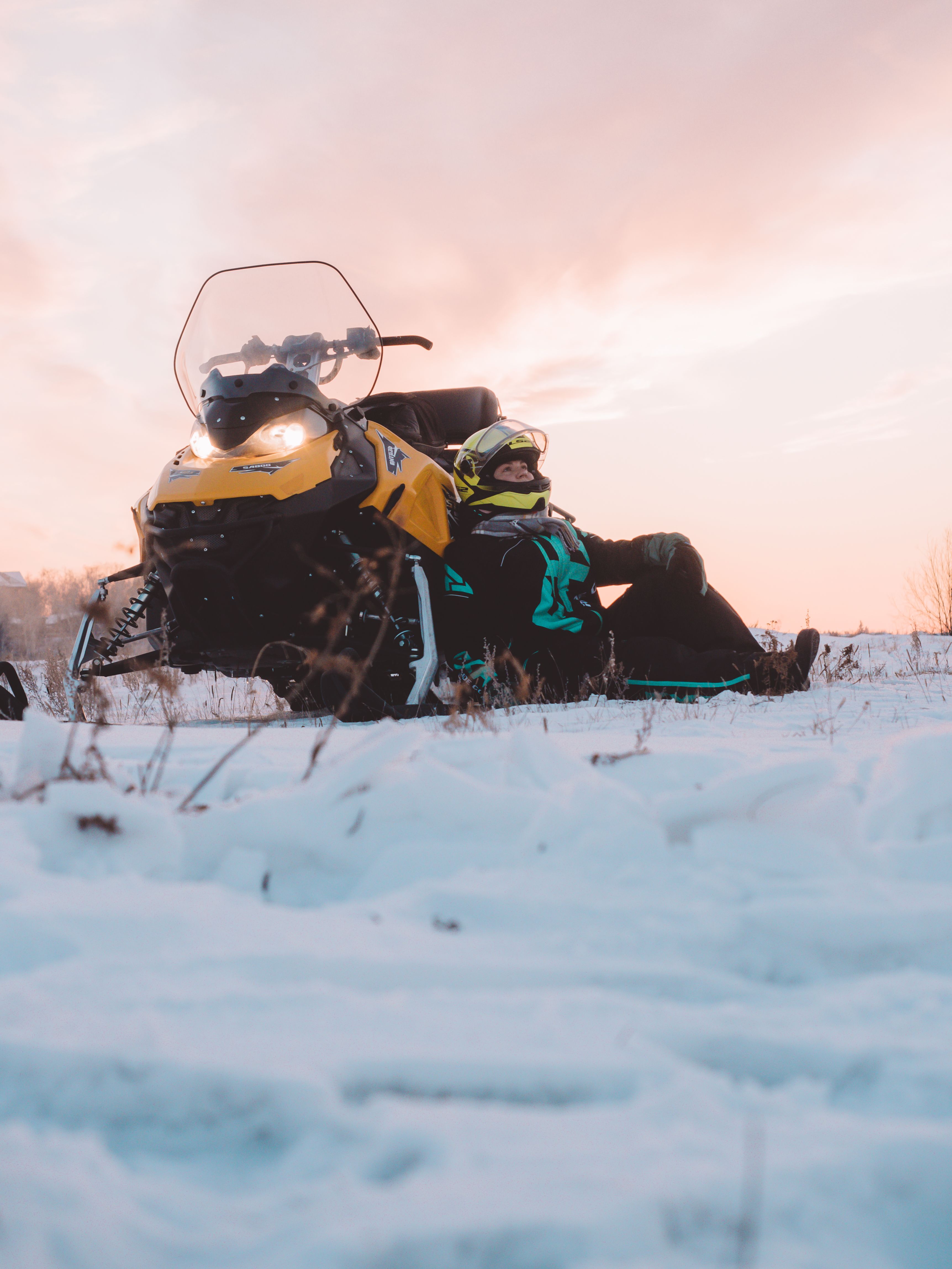 A person in winter gear sits beside a snowmobile on a snowy landscape at sunset.