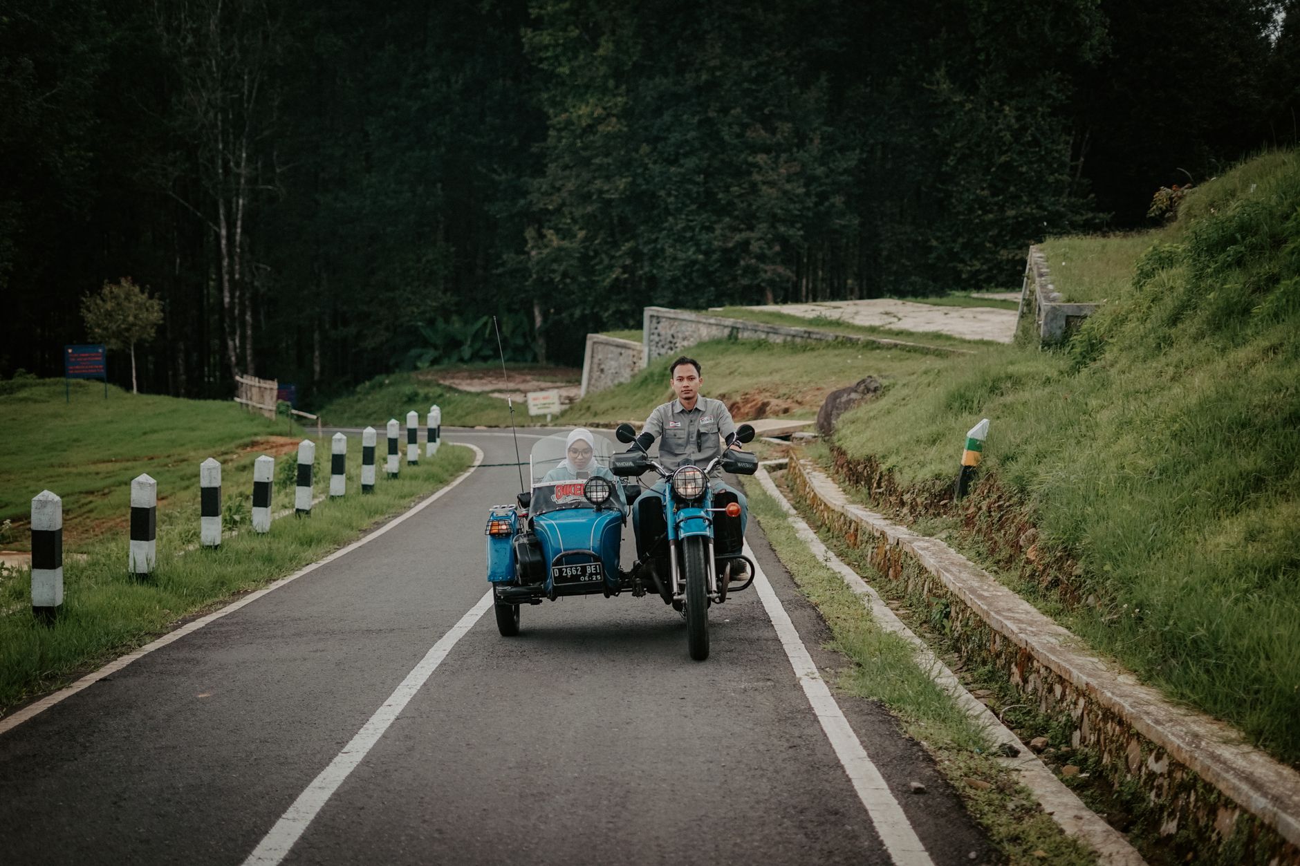 A couple riding a blue motorcycle with sidecar on a scenic, winding road through lush greenery.