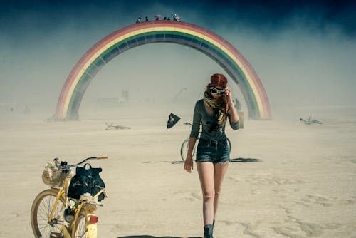 Woman in Motorcycle Goggles Walking in the Desert During a Sandstorm with a Rainbow Footbridge in the Background