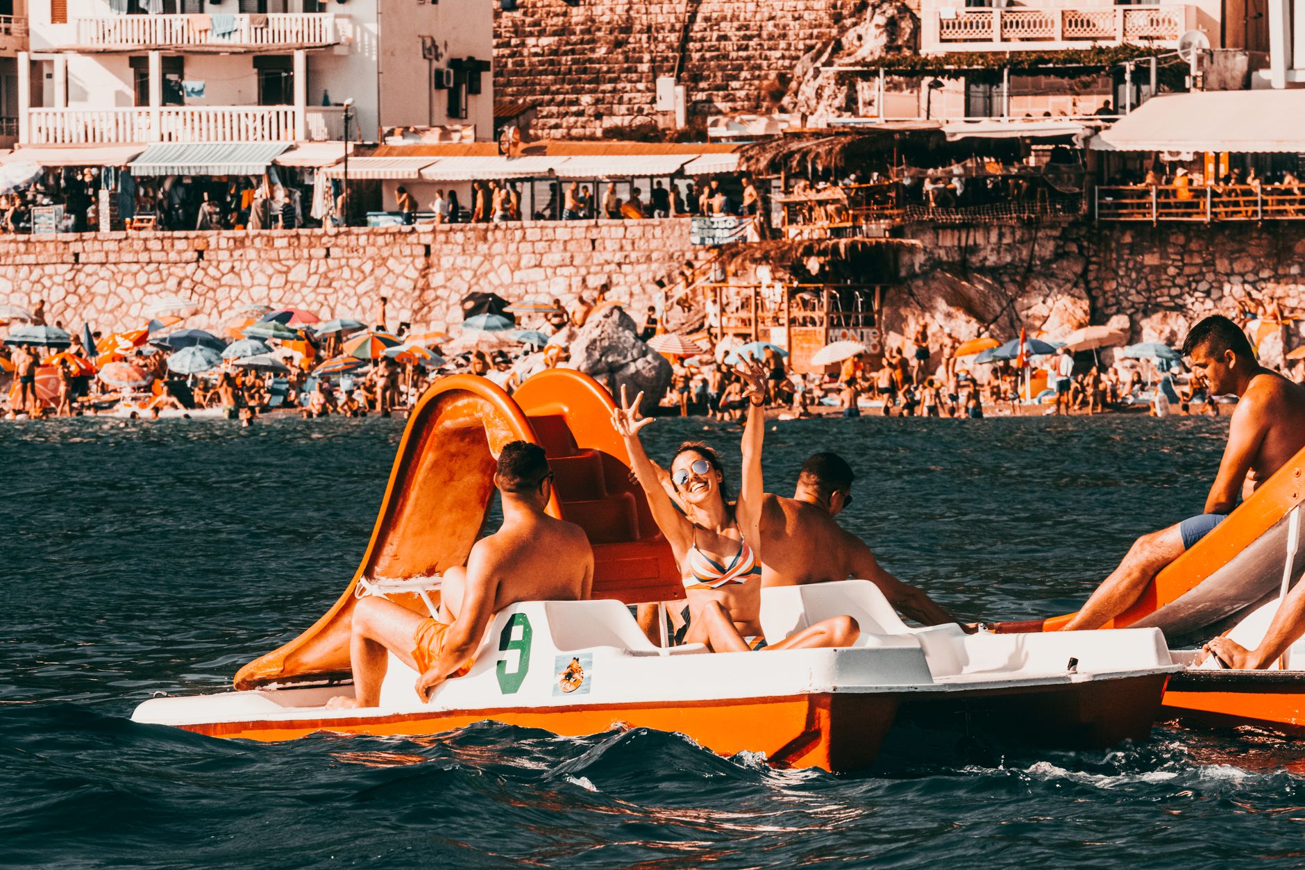 Tourists enjoying a sunny day riding pedalos on the coast of Montenegro with a lively beach in the background.