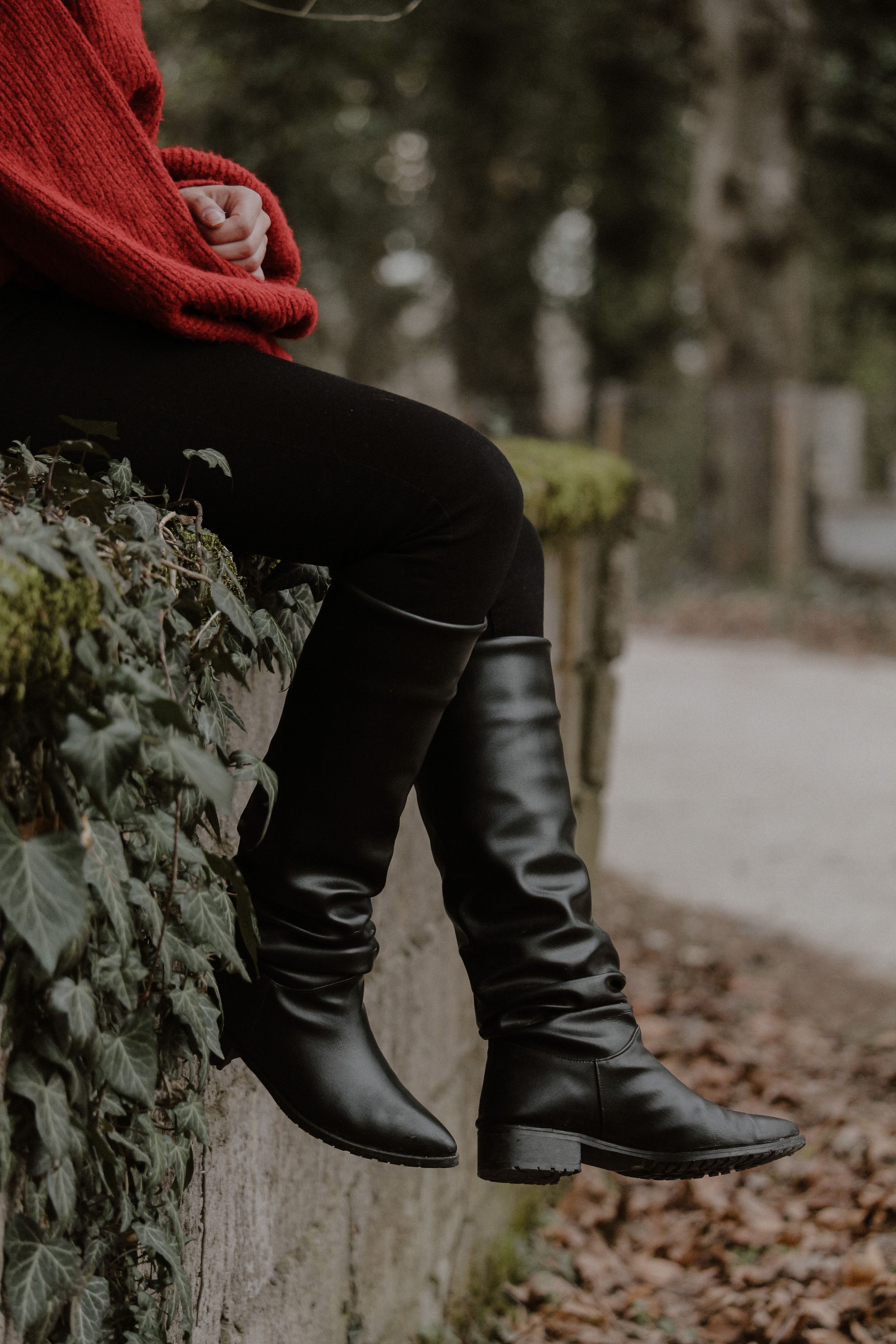 Uma pessoa com um suéter vermelho e botas pretas sentada em um muro coberto de musgo ao ar livre.