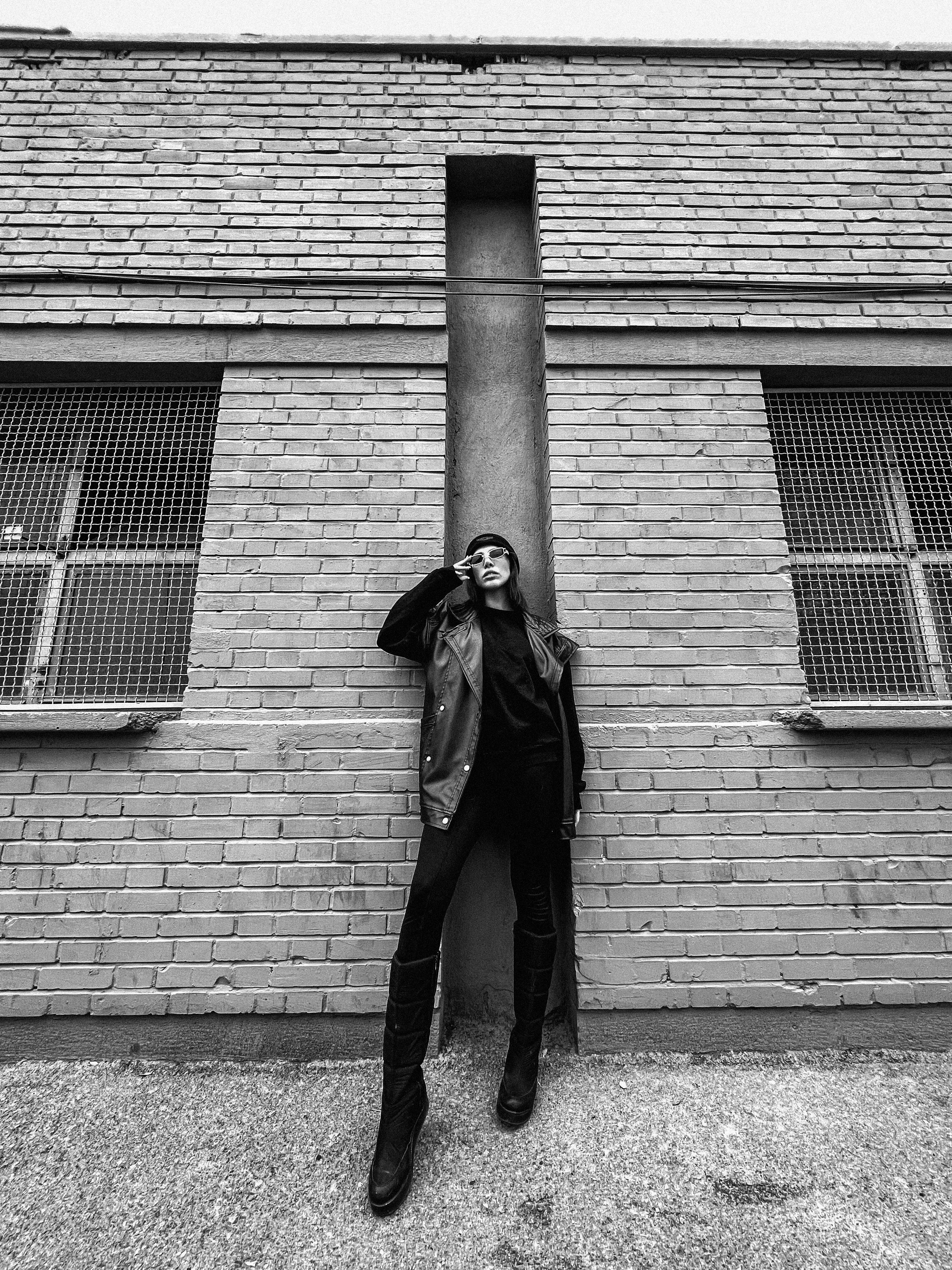 Black and white portrait of a woman in dark clothing leaning against a brick wall.