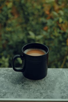 A black ceramic mug filled with coffee placed on a stone ledge with a blurred green background.