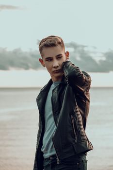 Young man posing fashionably in a black leather jacket by the sea during sunset.