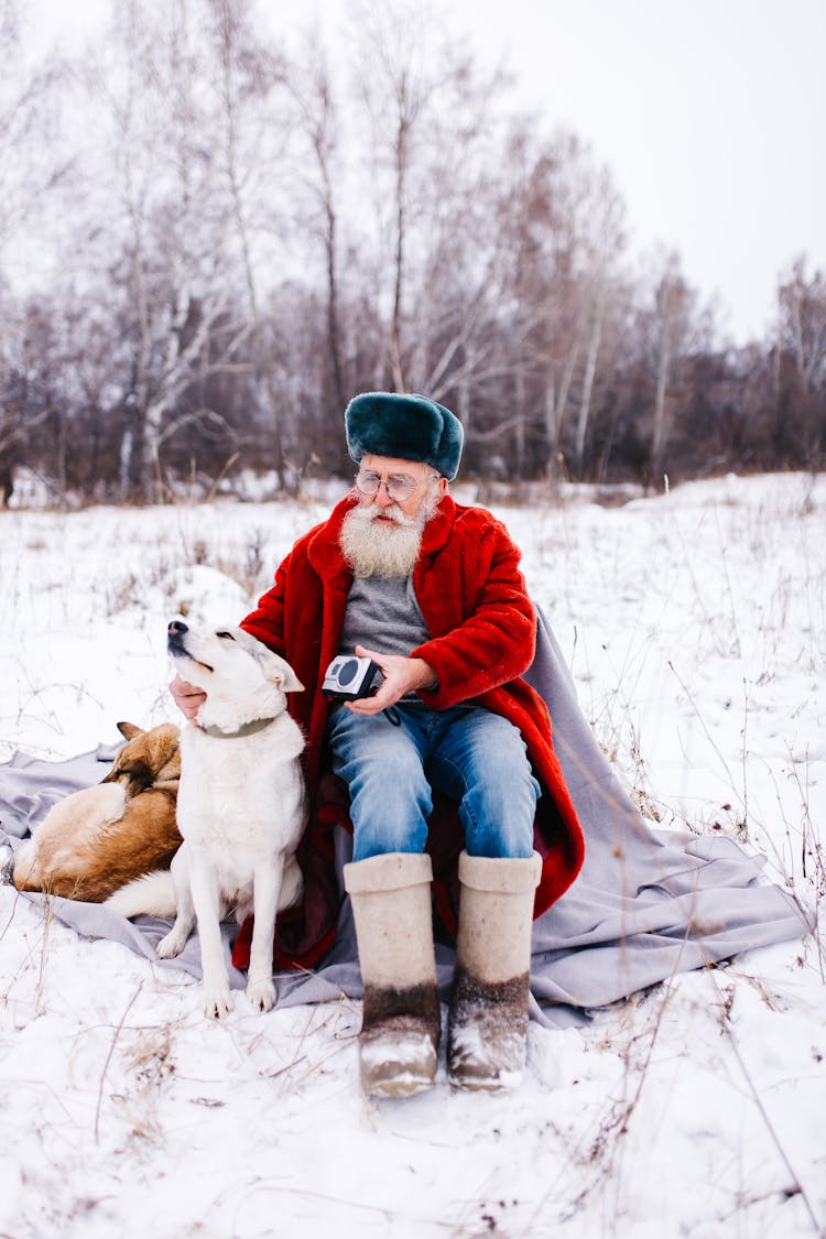 Elderly Man In Red Coat Sitting On Blanket In Snow With Dog