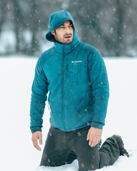 A man in a blue hoodie kneels in a snow-covered landscape during snowfall.