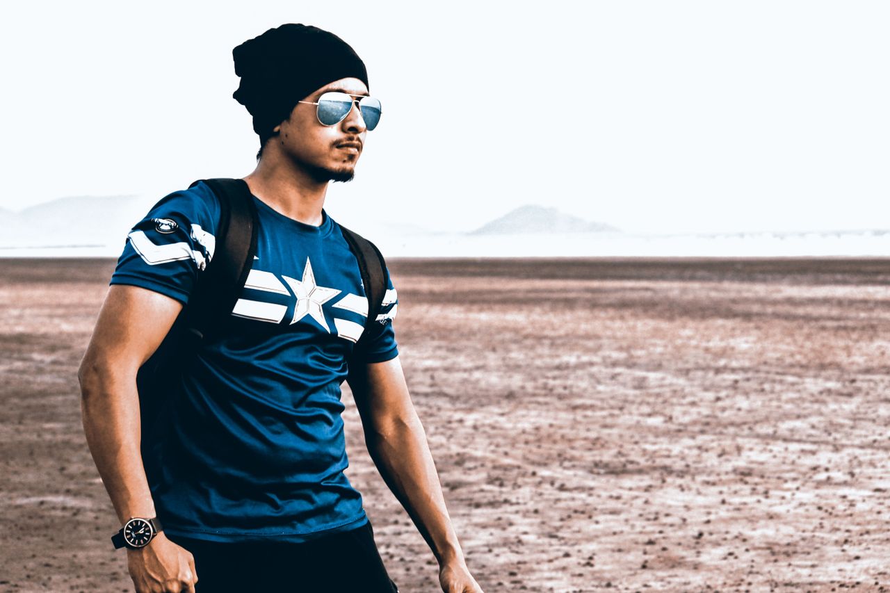 man in a beanie and sunglasses posing stylishly on a beach with mounta