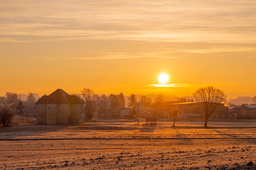 Peaceful townscape with warm orange sunrise over fields and trees, capturing serene nature and architecture.