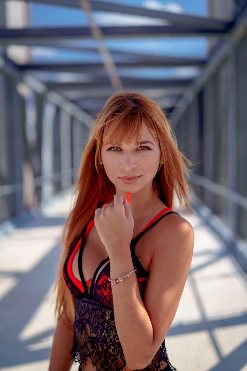 Free Smiling redhead woman in lace top posing outdoors under a metal structure. Stock Photo