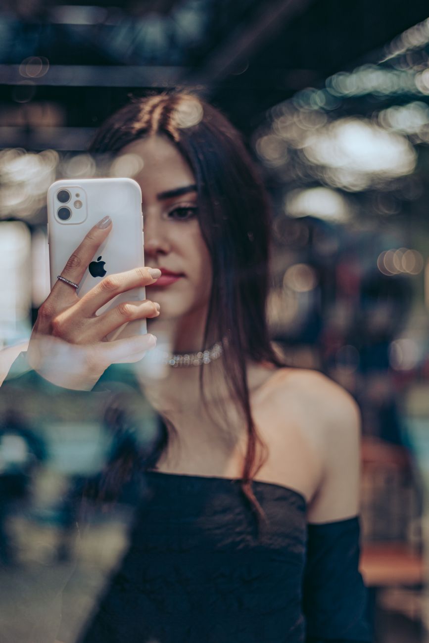 A fashionable woman takes a selfie with her smartphone, reflecting in the glass indoors.