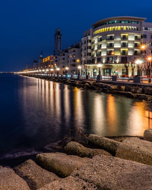 Free Beautiful nighttime cityscape featuring illuminated waterfront buildings along a calm shoreline. Stock Photo