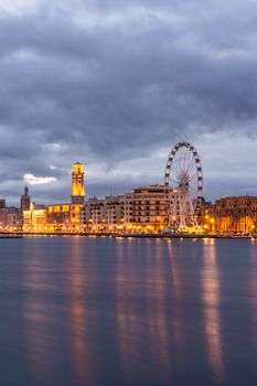 Illuminated waterfront cityscape with a Ferris wheel reflected in the river at twilight.