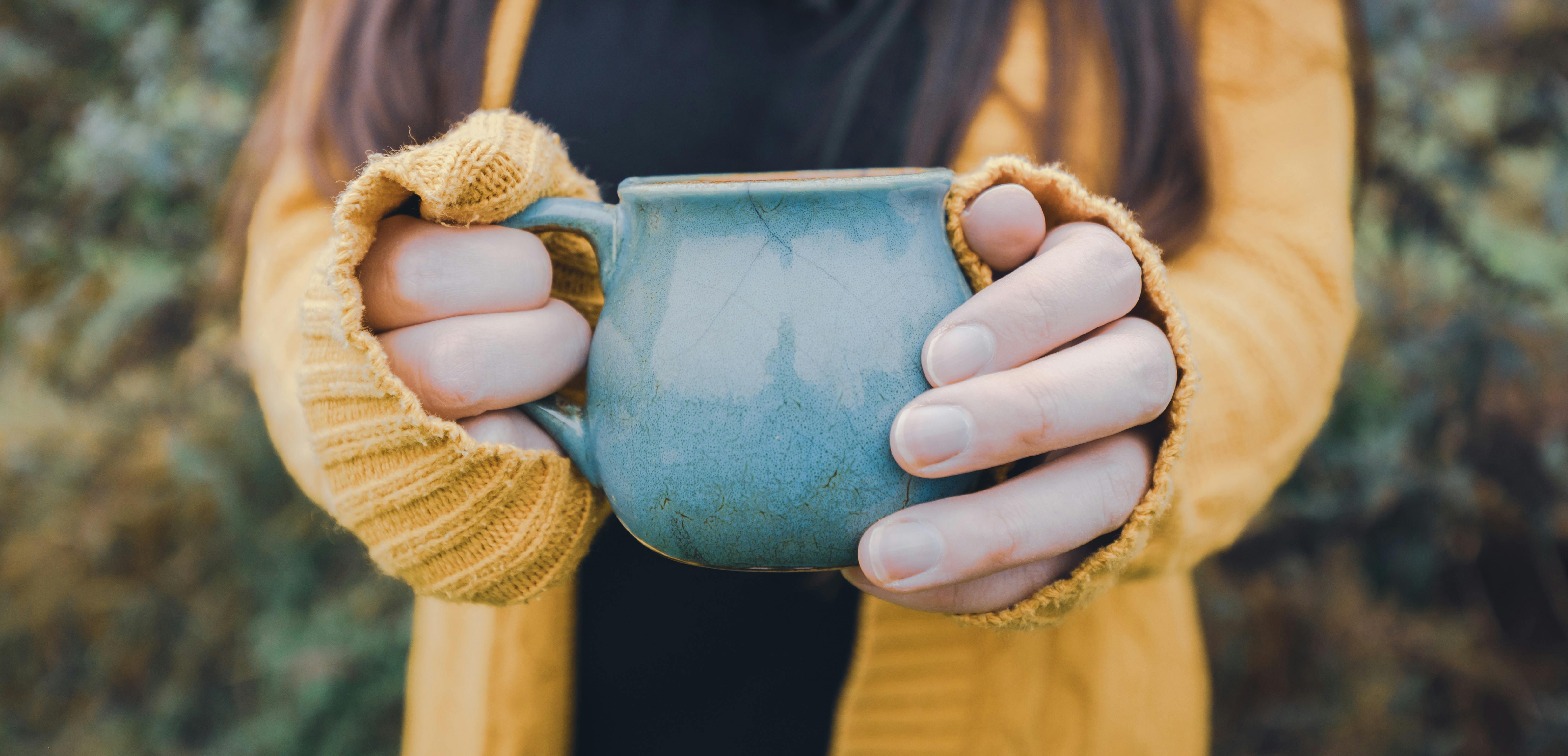 Close-up of hands in a cozy sweater holding a ceramic cup outdoors.