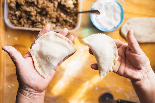 Close-up of hands preparing dumplings with flour and filling, bright kitchen setting.