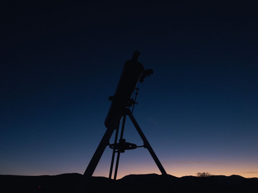 A telescope on a tripod in silhouette against a deep blue twilight sky
