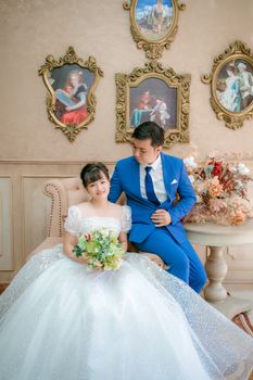A bride and groom in formal attire pose indoors amidst classic decor.