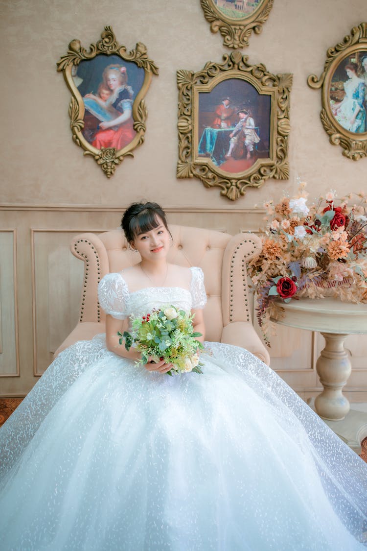 Woman In White Dress Holding Bouquet Of Flowers While Sitting On Beige Armchair Under Framed Pictures On The Wall